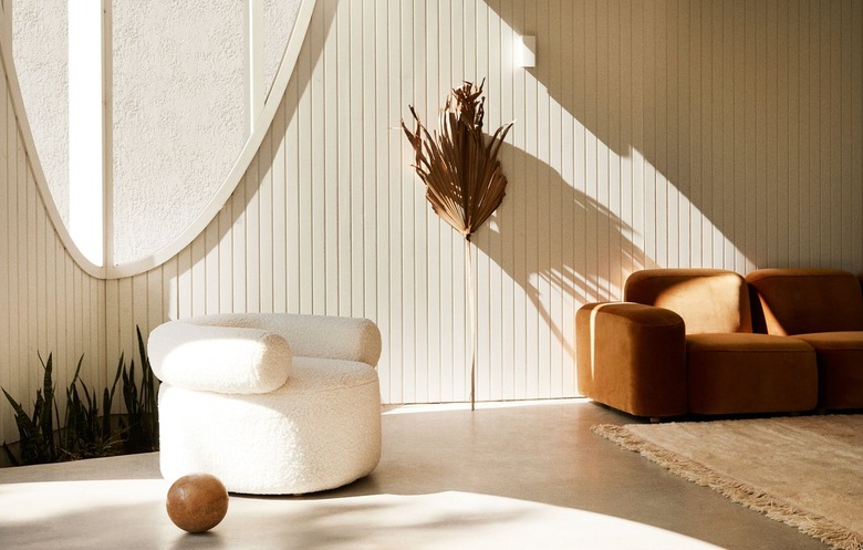 Minimalist living space with natural sunlight from large, uniquely shaped windows. A plush white shearling armchair, a section of a terracotta velvet sofa, a braided rug, a wooden ball, and a tall, dried palm leaf are visible. The wall has vertical wood paneling, and plants are in a lower area.